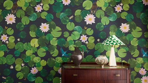 Wallpaper with a bold water lilly print in deep greens and pink sits behind a sideboard with a lamp and vases on top.